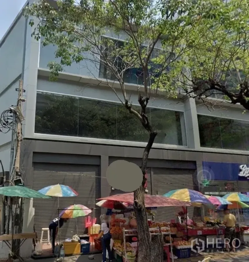 Retail Space for rent in Watthana, Bangkok - Street-facing modern retail building with large glass windows, roll-up shutters, and colorful market umbrellas under trees in Watthana, Bangkok.