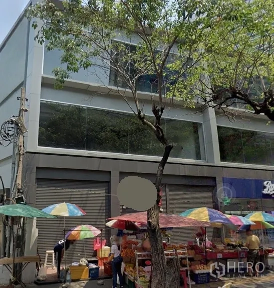 Retail Space for rent in Watthana, Bangkok - Street-facing modern retail building with large glass windows, roll-up shutters, and colorful market umbrellas under trees in Watthana, Bangkok.