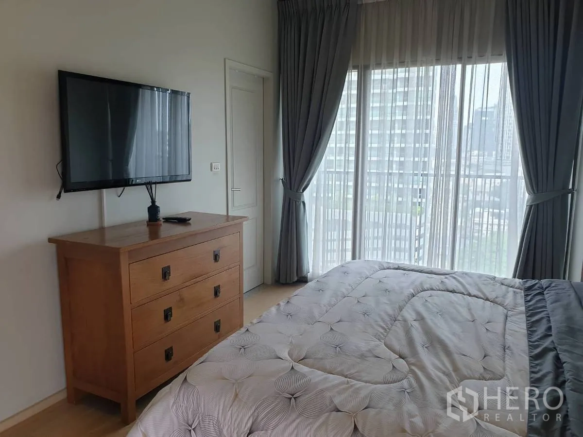Condominium for rent or sale in Bangkok - Bedroom with wall-mounted TV and floor-to-ceiling sheer curtains overlooking the city.