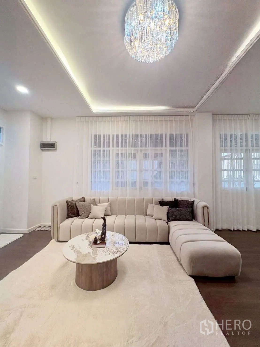 Townhouse for rent in Huai Khwang, Bangkok - Living room with large L-shaped sofa, round marble-top table, and crystal light.