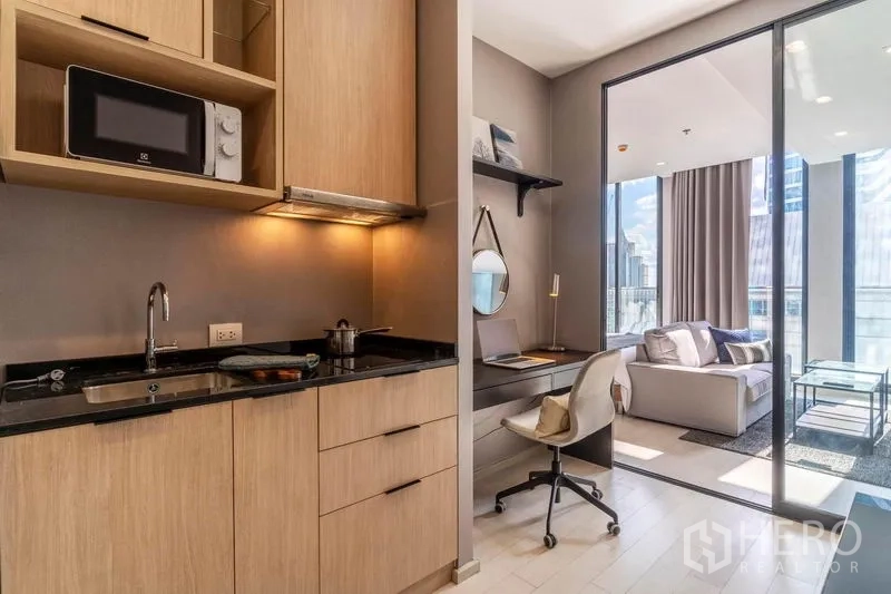 Condominium for rent in Pathum Wan, Bangkok - Compact kitchenette with wood cabinetry and black countertop beside a built-in study desk, linked to the lounge by sliding glass.
