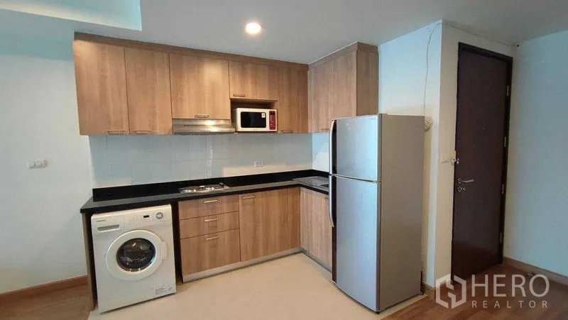 Condominium for rent in Khlong Toei, Bangkok - Fitted kitchenette with wood cabinets, black countertop, fridge, microwave and washing machine.