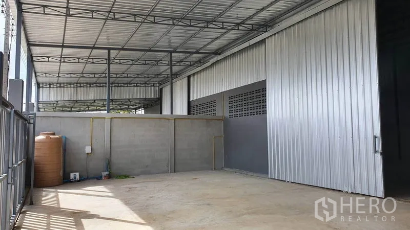 Warehouse for rent in Lam Luk Ka, Pathum Thani - Side service yard with water tank, utility lines and corrugated metal walls beneath canopy.