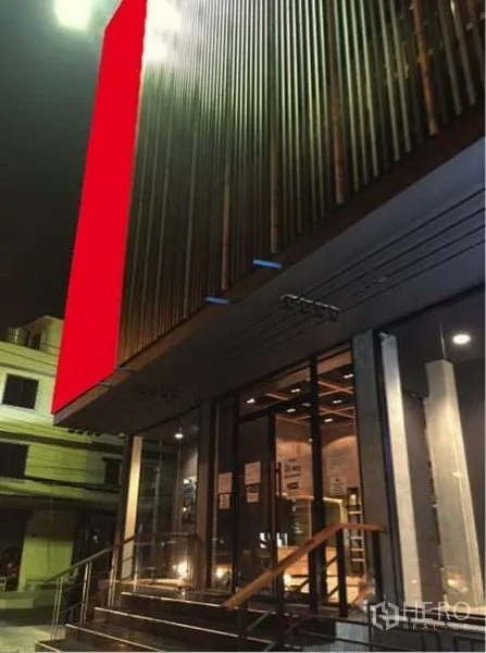 Shophouse for rent in Suan Luang, Bangkok - Nighttime exterior of a modern shophouse with glass storefront, metal slat facade, large red sign panel, and steps with railings.