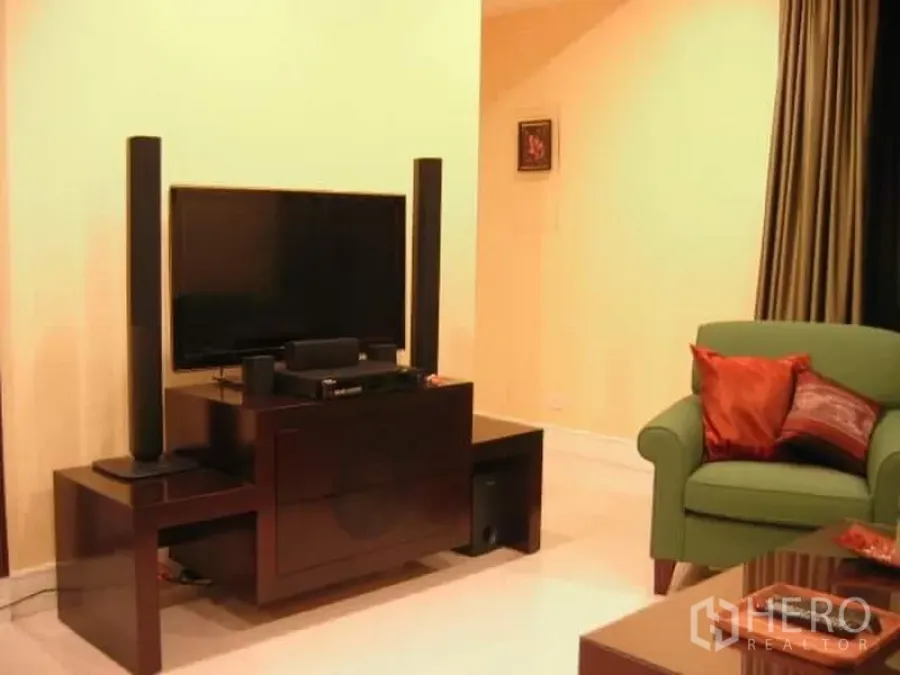 Condominium for sale in Khlong Toei, Bangkok - Warm-toned living room with flat-screen TV on dark wood console, tall speakers, green armchair and full-length curtains.