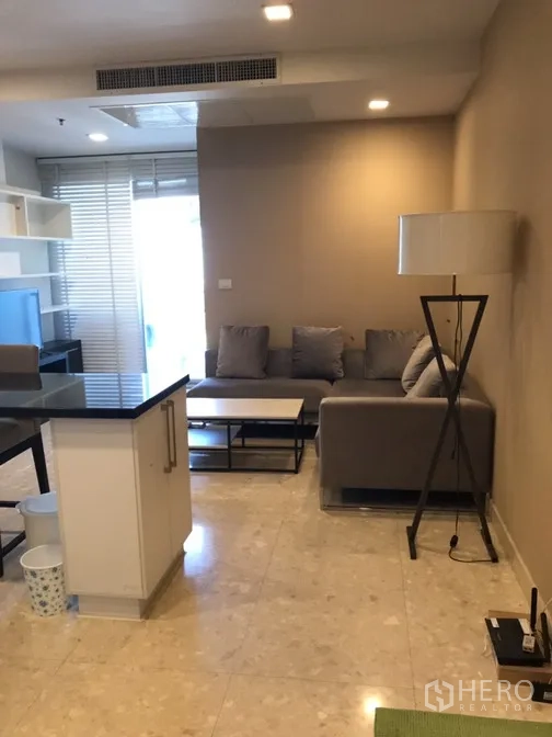 Condominium for rent in Khlong Toei, Bangkok - Open-plan lounge with sectional sofa, floor lamp and sliding door to a balcony beside the kitchen island.