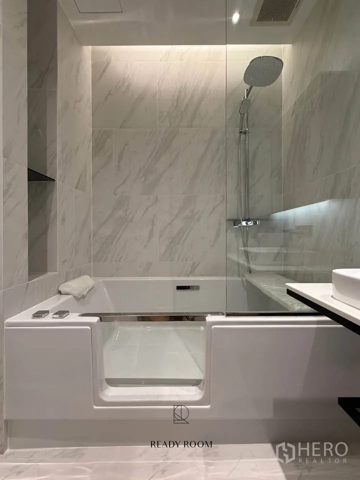 Condominium for rent in Khlong Toei, Bangkok - Marble-tiled bathroom with glass shower, rain head, and soaking tub with a side door.