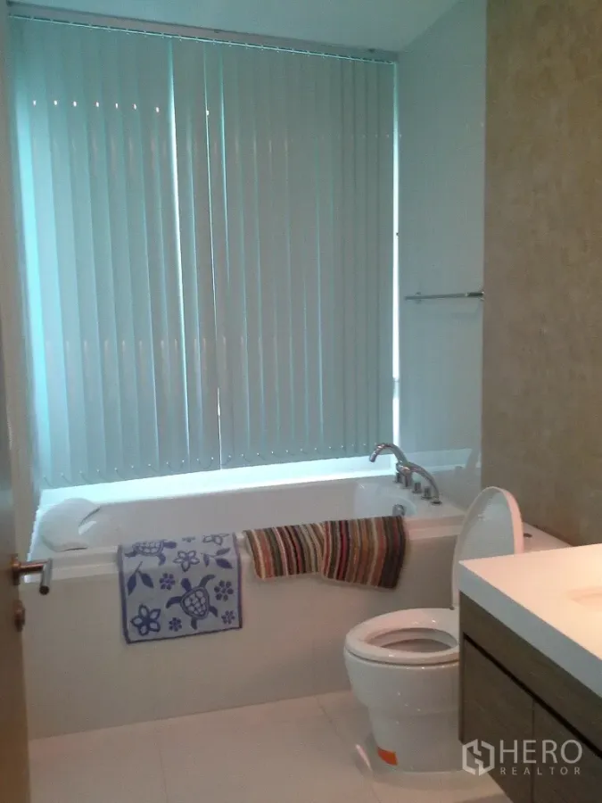 Condominium for rent or sale in Khlong Toei, Bangkok - Bright bathroom with a bathtub, vertical blinds, toilet, and vanity on white tiles.