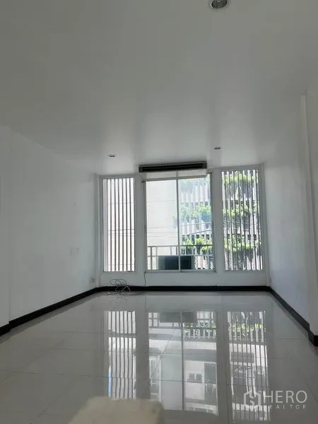 Shophouse for rent in Khlong Toei, Bangkok - Bright upper‑floor room with white tile flooring, tall windows and a sliding door to a small balcony.