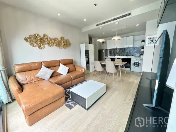 Condominium for rent in Bangkok - Open-plan lounge with L-shaped tan sofa, dining table and compact kitchen beside floor-to-ceiling windows.