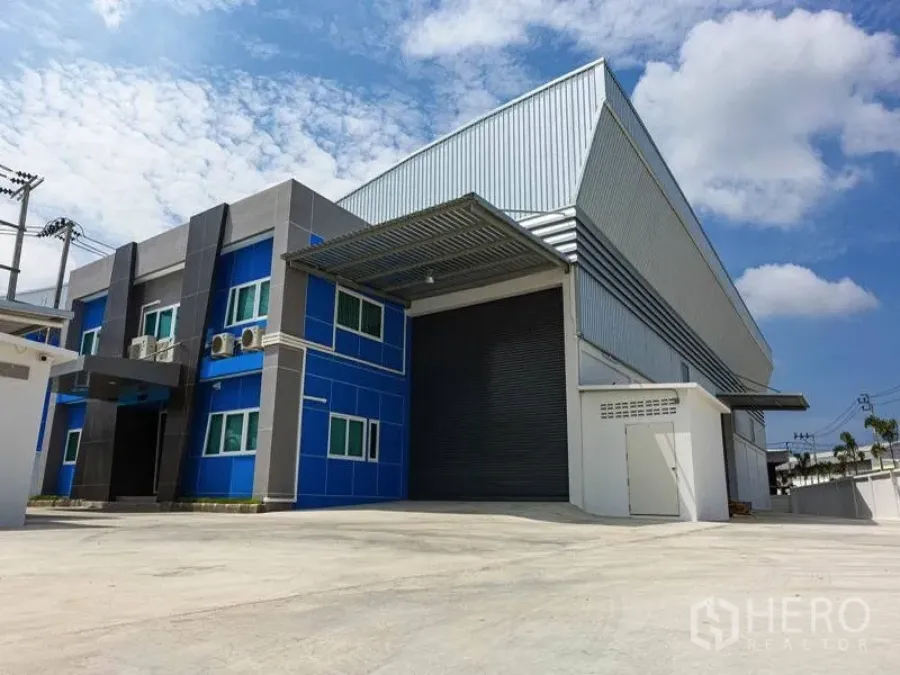Factory for rent in Bang Bo, Samut Prakan - Modern factory exterior in Bang Bo with blue office wing, large roller shutter, covered loading bay and spacious concrete yard under a clear sky.
