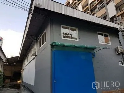 Warehouse for rent in Chatuchak, Bangkok - Gray warehouse exterior in Chatuchak with blue steel sliding door, small windows, AC units and corrugated metal roof beside mid‑rise buildings.