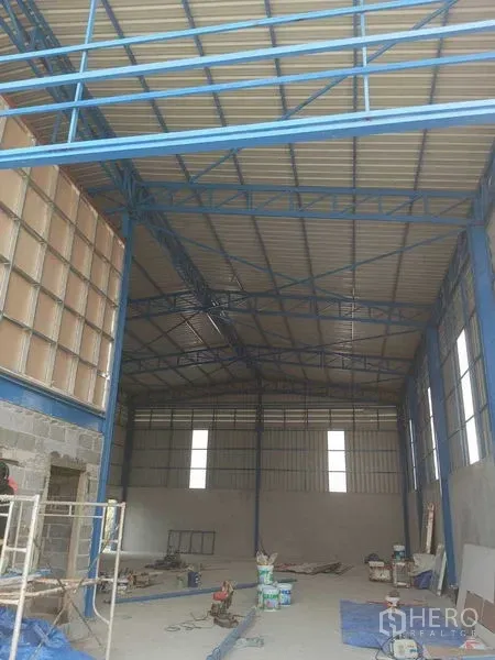 Warehouse for rent in Pathum Thani - Spacious warehouse interior showing high-clearance roof, blue steel trusses, and construction materials on the floor.