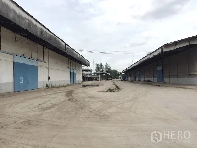 Warehouse for rent in Pathum Thani - Spacious truck yard between two long concrete warehouses with blue sliding doors in Pathum Thani.