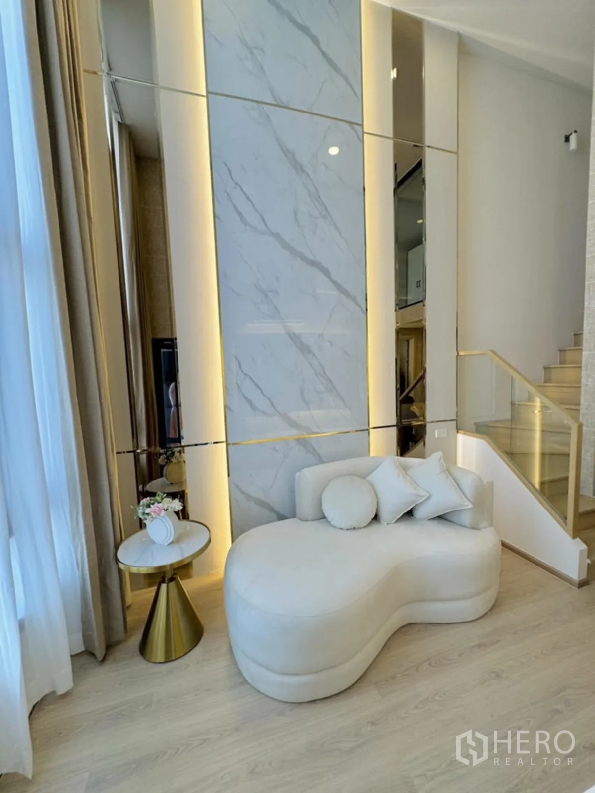Condominium for rent in Khlong Toei, Bangkok - Bright living area with cream chaise lounge, marble feature wall with warm lighting, large curtains, light wood floors, and a glass-railed staircase.