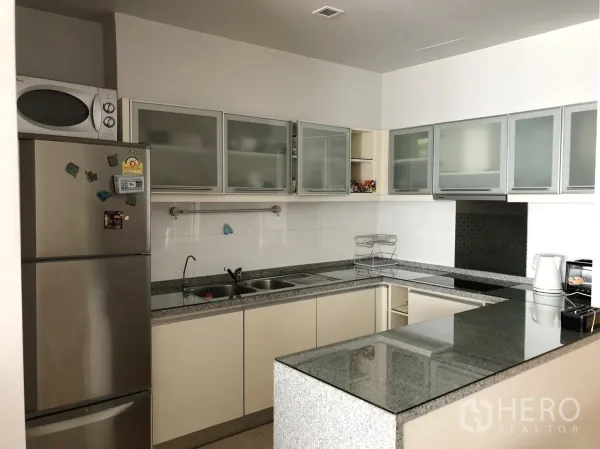 Condominium for rent or sale in Khlong Toei, Bangkok - U-shaped kitchen with granite countertops, glass-front cabinets, stainless refrigerator and microwave.