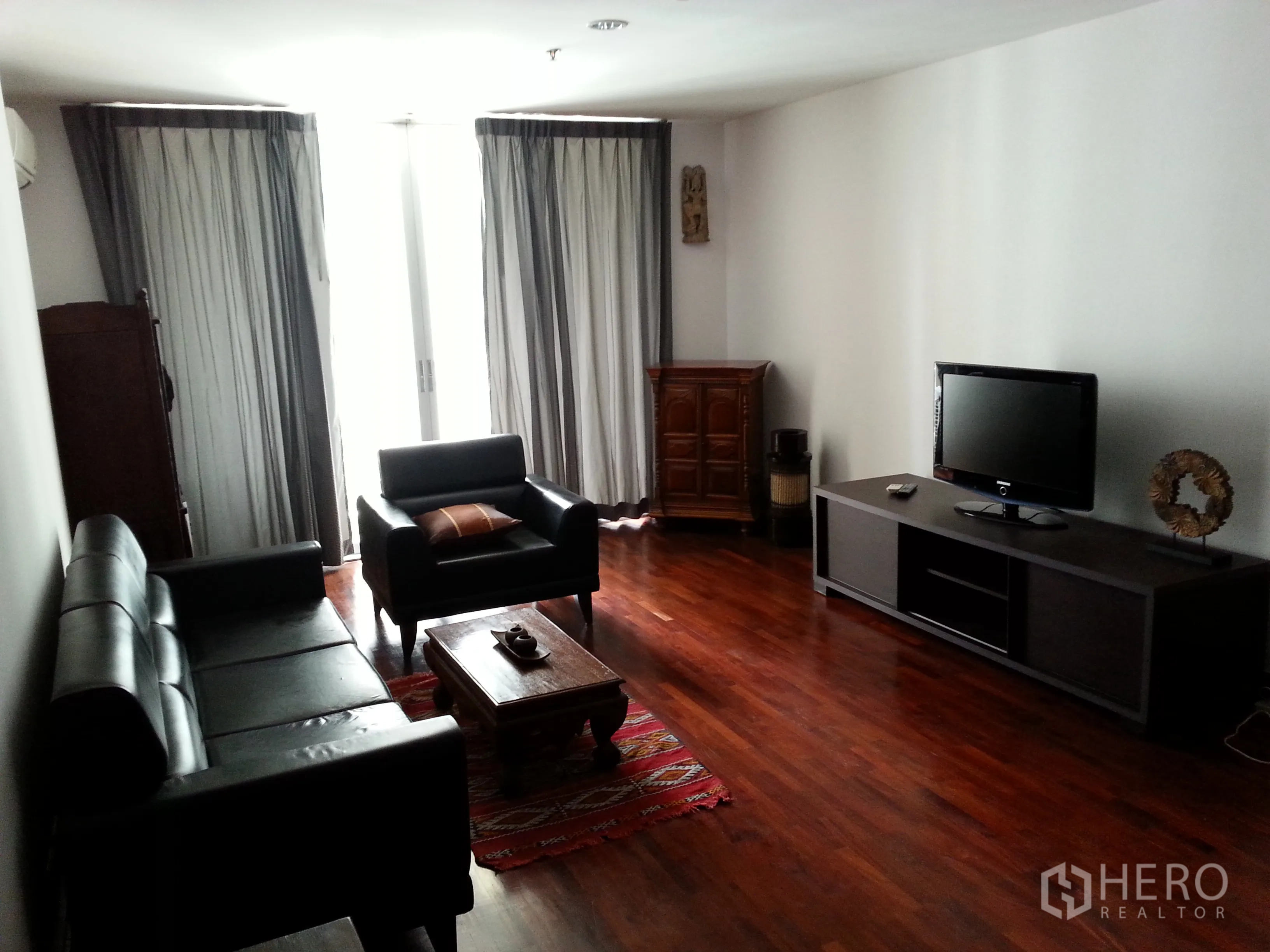 Condominium for rent in Watthana, Bangkok - Bright condo living room with floor-to-ceiling curtains, dark leather sofa set, hardwood floors, TV console and wooden cabinet.