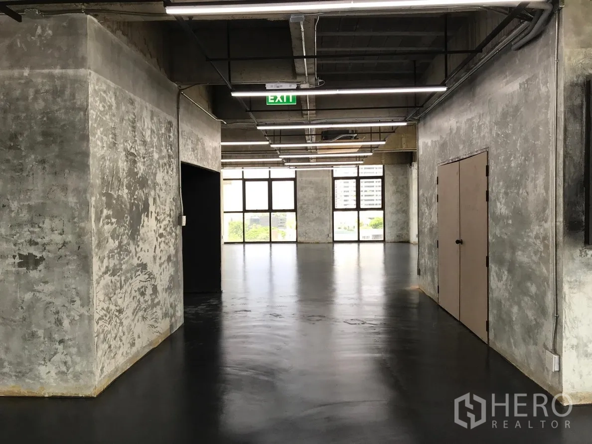 Office Space for rent in Khlong Toei, Bangkok - Hallway view of the office level with textured concrete walls, emergency exit sign, double utility doors, and daylight from end windows.