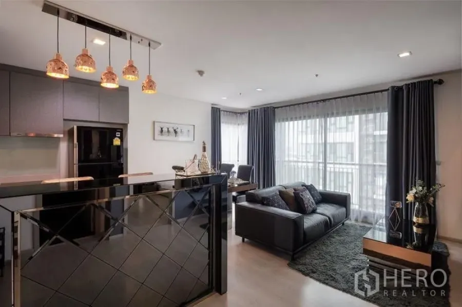 Condominium for rent or sale in Khlong Toei, Bangkok - Open-plan living and kitchen with pendant lights, black sofa, and floor-to-ceiling windows.
