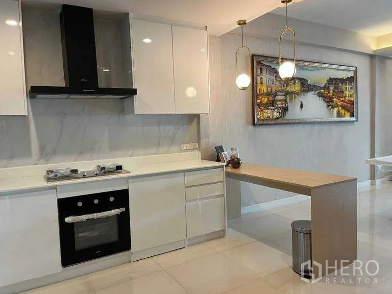 Townhouse for rent in Khlong Toei, Bangkok - Modern white kitchen with marble backsplash, built-in oven, hob, and breakfast bar under pendant lights.