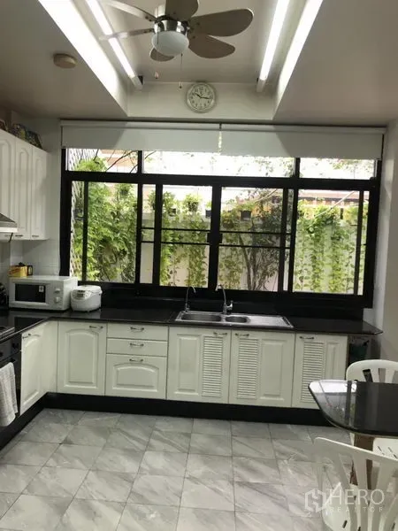 Townhouse for rent or sale in Bangkok - Bright kitchen with white cabinets, black countertops, large window to green vines, and a ceiling fan.