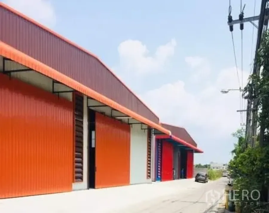 Warehouse for rent in Bueng Kum, Bangkok - Row of modern metal-clad warehouse units with bold orange façades and a wide driveway for truck access.