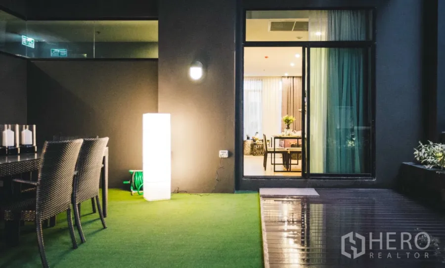 Condominium for rent in Huai Khwang, Bangkok - Private terrace with artificial grass, outdoor dining set, and sliding door opening to a bright living area at night.