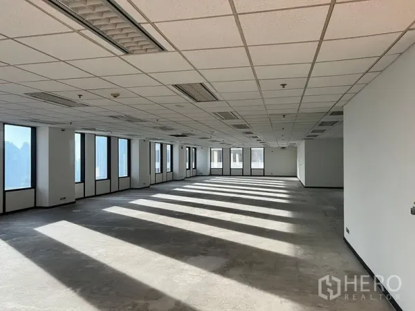 Office Space for rent in Khlong Toei, Bangkok - Sunlit open‑plan office floor with drop ceiling, fluorescent lights, and long window shadows across a bare concrete floor, with multiple windows showing city views.