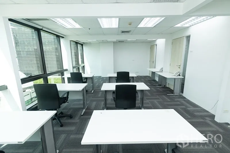 Office Space for rent - Open office area with arranged desks and chairs beside full-height windows and clean white walls.