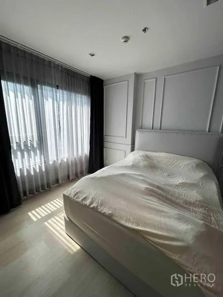 Condominium for sale in Pathum Wan, Bangkok - Bright bedroom with upholstered bed, floor‑to‑ceiling window, sheer and blackout curtains, and elegant wall paneling.