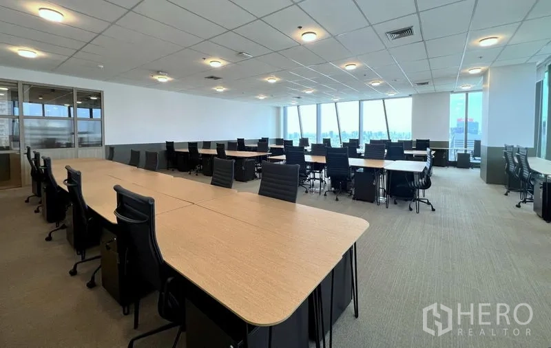 Office Space for rent - Open-plan office with long workstations, ergonomic chairs, carpet tiles, acoustic ceiling lights, and floor-to-ceiling windows showing Bangkok skyline views.