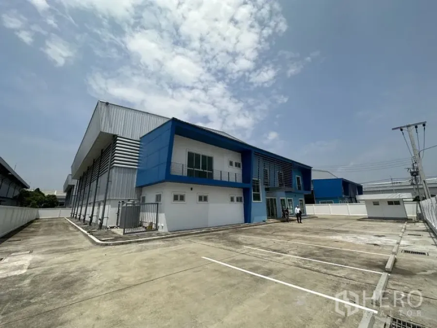 工厂 出租在Bang Sao Thong, Samut Prakan - 蓝白色现代独栋工厂，带两层办公楼、围栏及宽阔混凝土停车场，天空晴朗。