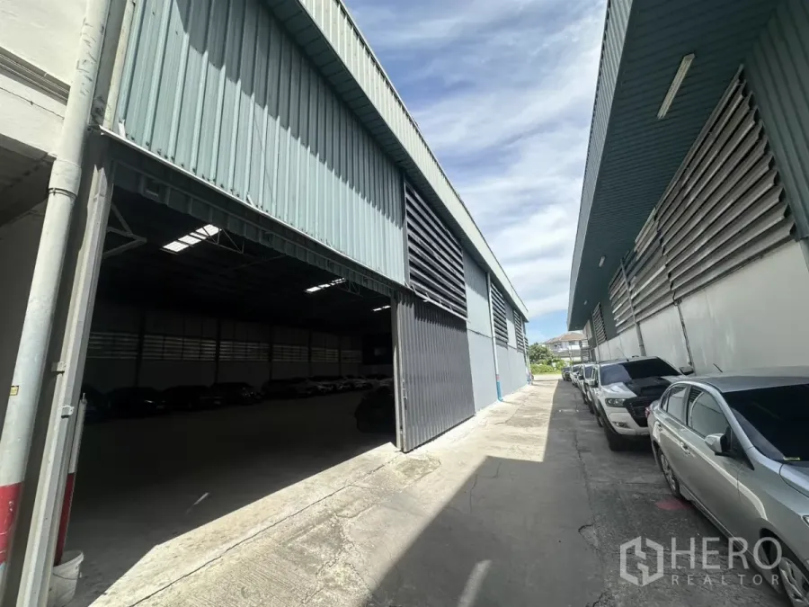Warehouse for rent in Suan Luang, Bangkok - Exterior view of a metal warehouse with a wide sliding door open and a side driveway with parked cars.