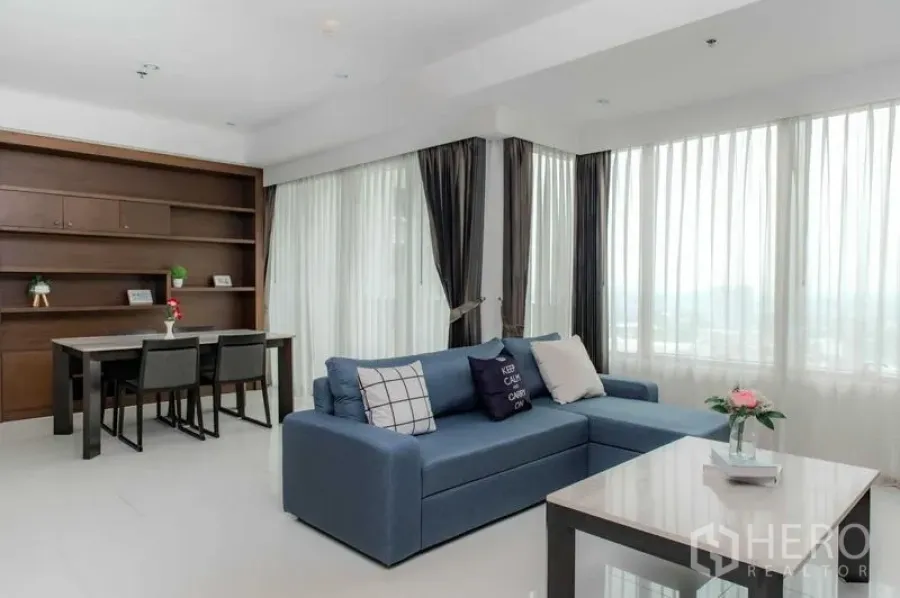 Condominium for rent or sale in Bangkok - Living and dining room with blue L-shaped sofa and bright windows.