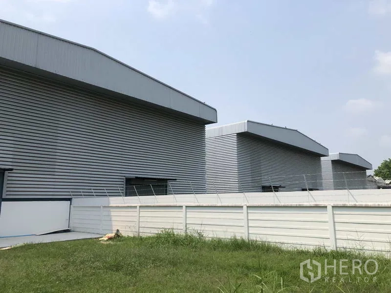Factory for sale in Sai Noi, Nonthaburi - Row of modern factory buildings behind a white perimeter fence bordering a grassy yard.