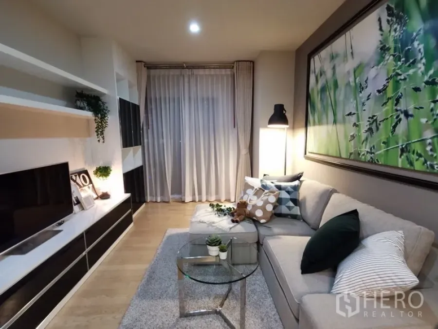 Condominium for sale in Bangkok - Cozy living room featuring an L-shaped sofa, built-in TV wall, sheer drapes, and soft ambient lighting.