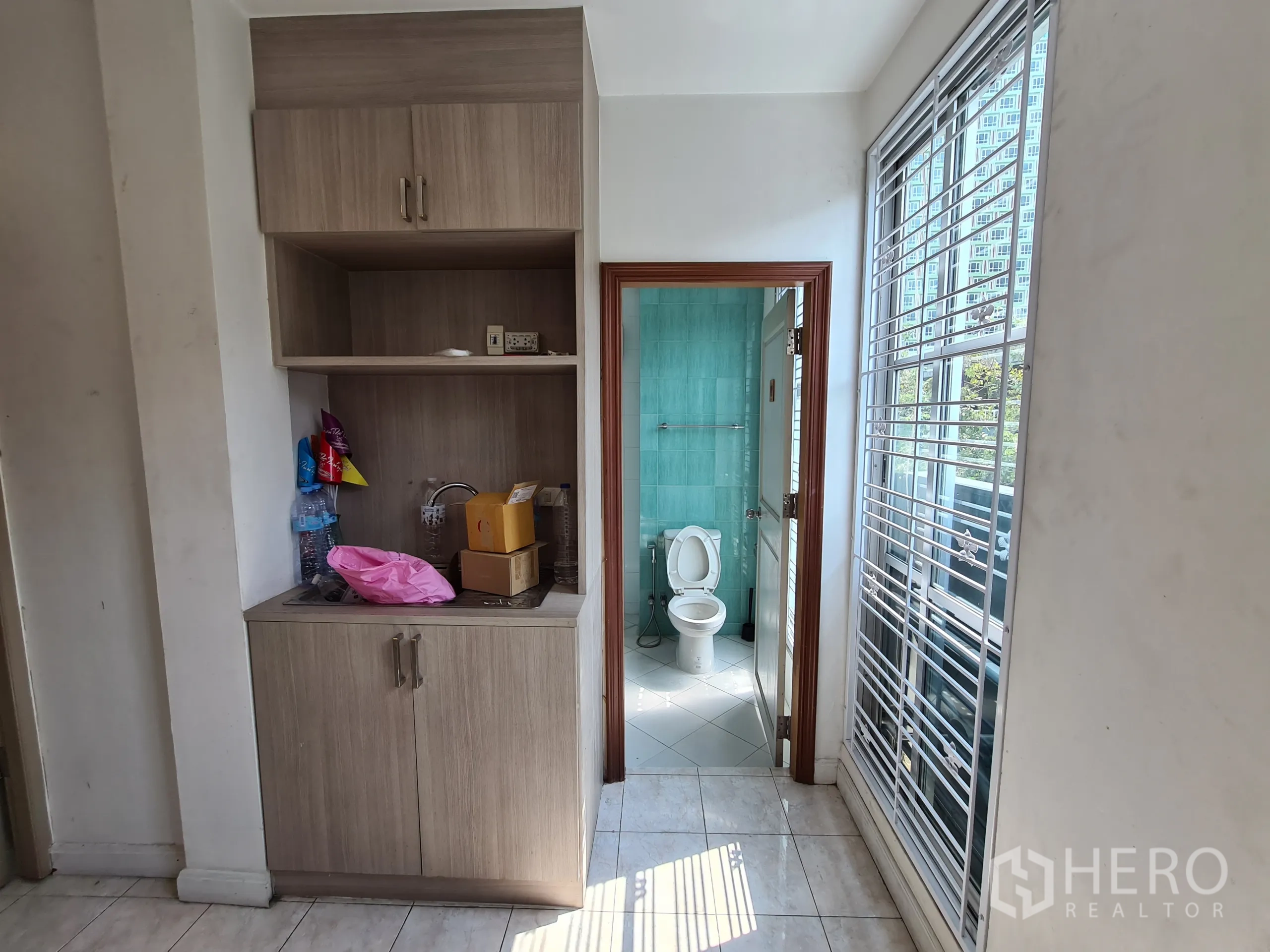 Shophouse for rent or sale in Sathon, Bangkok - Compact kitchenette with cabinets beside a tiled bathroom and tall grilled window.