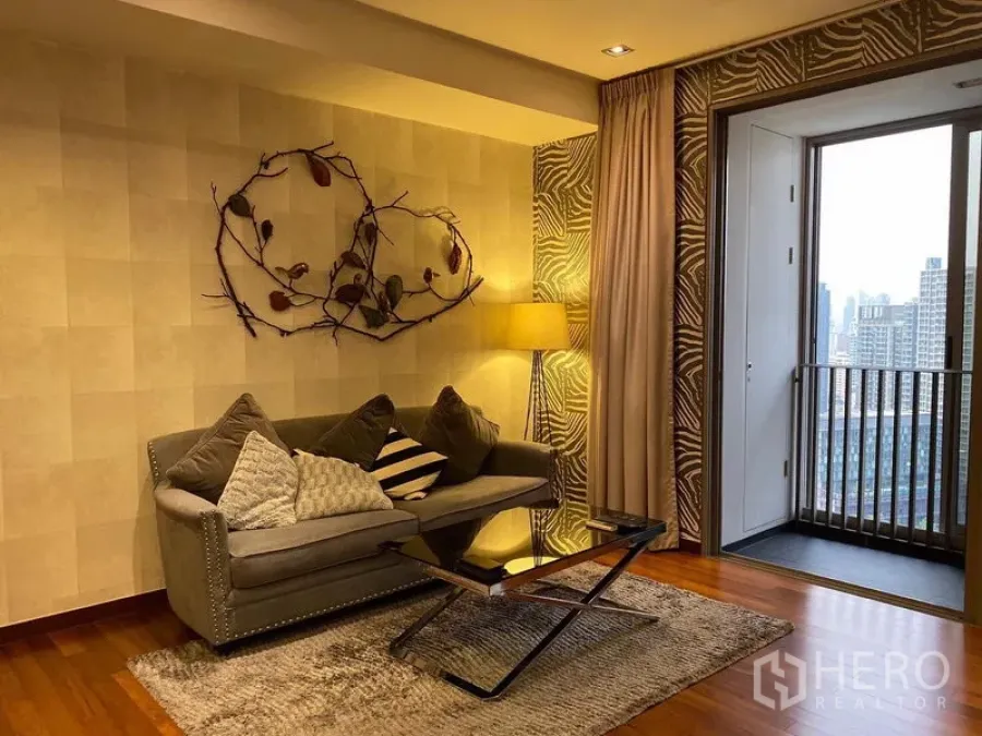 Condominium for rent in Khlong Toei, Bangkok - Cozy living room with gray sofa, wall art, floor lamp, hardwood floors and a balcony city view.
