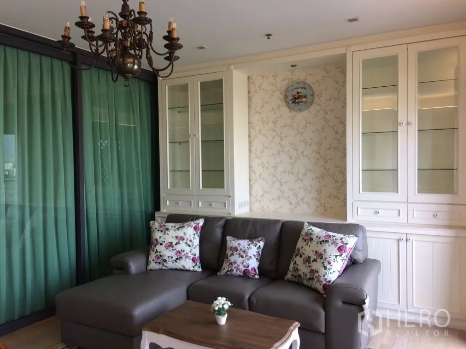 Condominium for rent in Khlong Toei, Bangkok - Cozy living room with gray sectional sofa, floral pillows, vintage chandelier and white built-in glass cabinets beside green drapes.