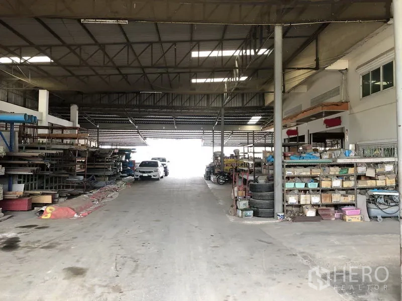 Warehouse for rent in Sai Noi, Nonthaburi - Drive-through central aisle with vehicles and shelving under a high steel roof and skylights.