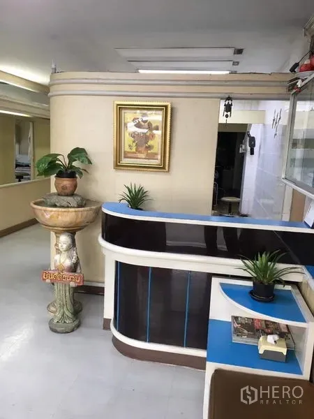 Shophouse for rent in Phra Khanong, Bangkok - Curved reception counter with potted plants and a decorative fountain against cream walls.