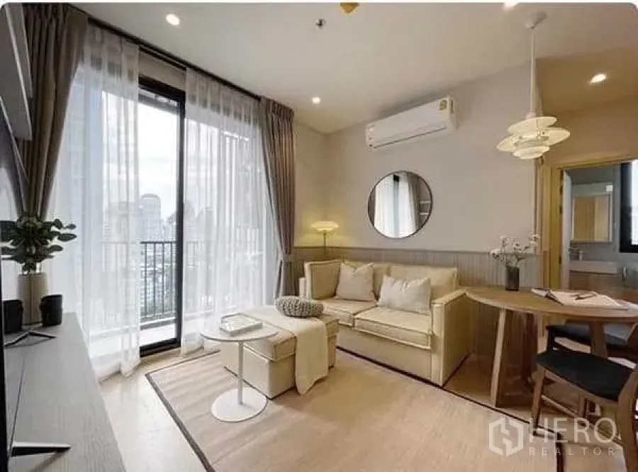 Condominium for rent in Watthana, Bangkok - Bright living room with beige sofa, round mirror, air conditioner and balcony city view near floor‑to‑ceiling windows.