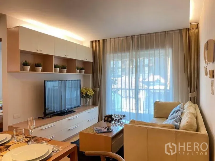 Condominium for rent or sale in Phra Khanong, Bangkok - Living room with sofa and TV on built‑in console facing sliding glass door to balcony.