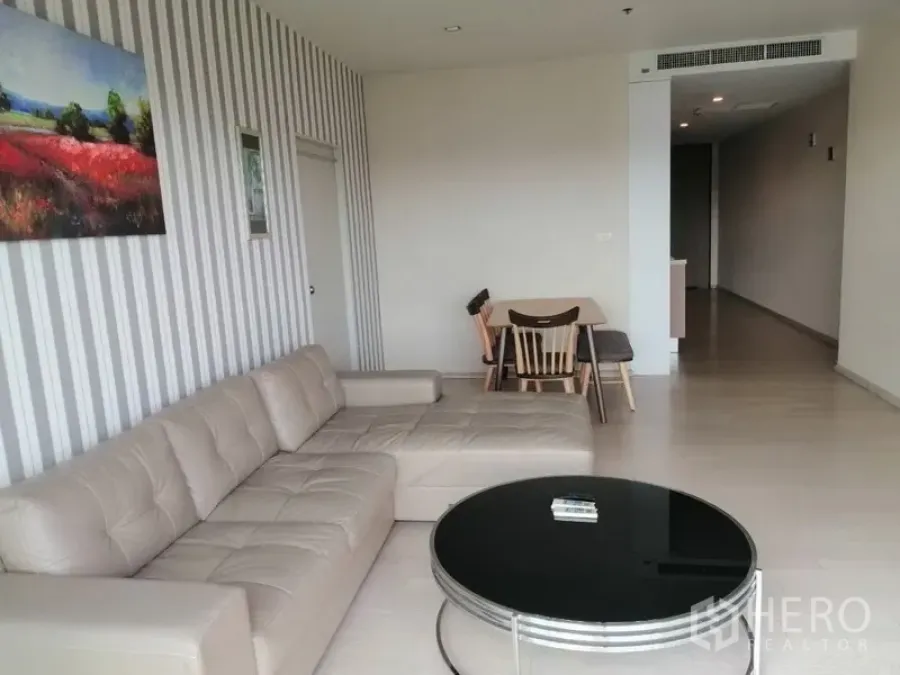 Condominium for rent in Watthana, Bangkok - Bright living room with beige L-shaped sofa, round coffee table, striped feature wall and a small dining set.