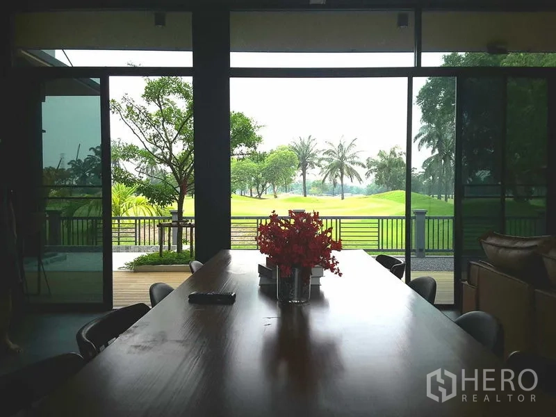 Detached House for rent or sale in Lam Luk Ka, Pathum Thani - Dining area with long wooden table and full-height windows looking out to garden and fairway.