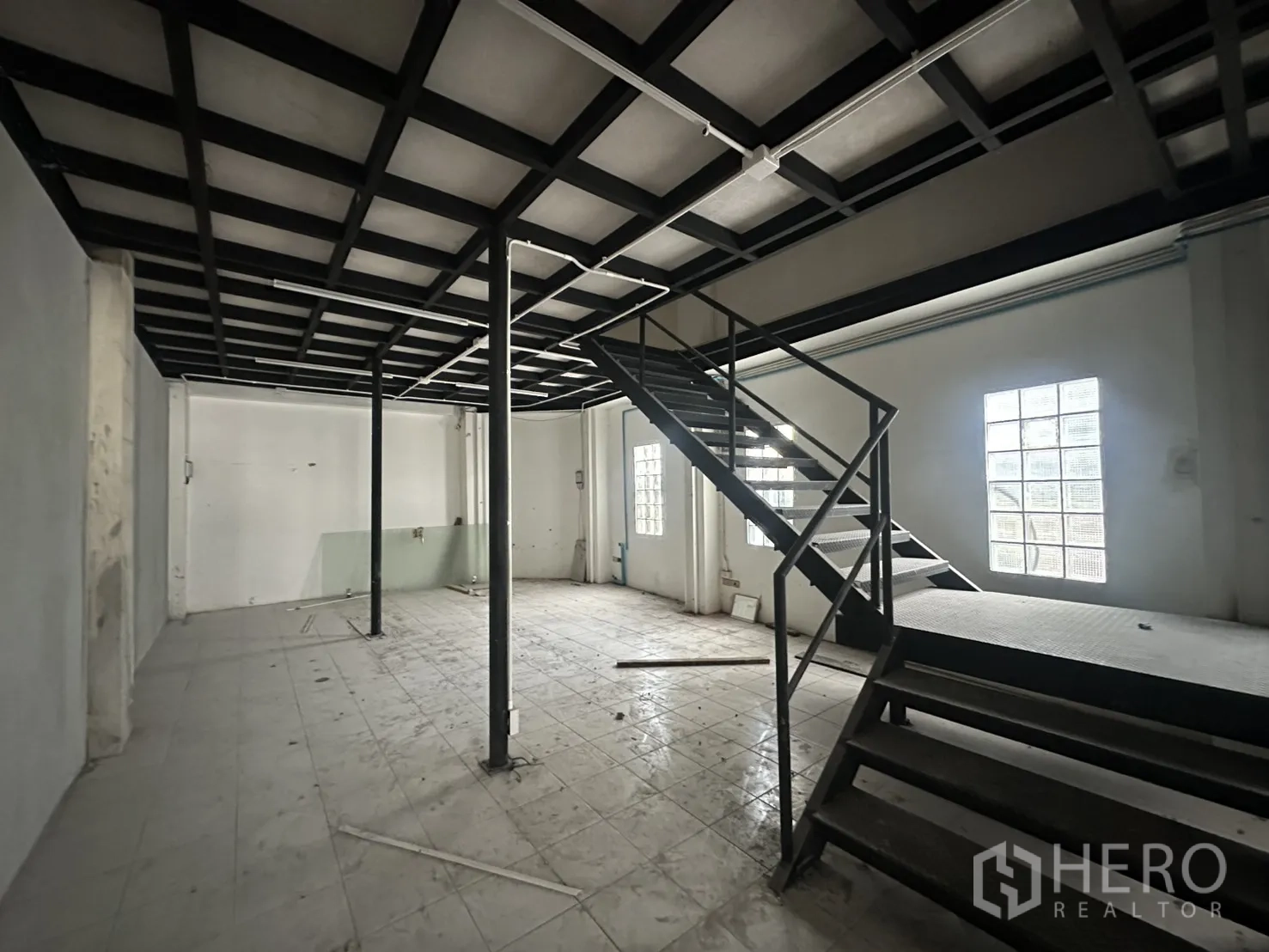 零售空间 for rent in Watthana, Bangkok - 开放式零售室内，瓷砖地面与黑色钢柱，金属楼梯通往夹层，旁有玻璃砖窗采光。
