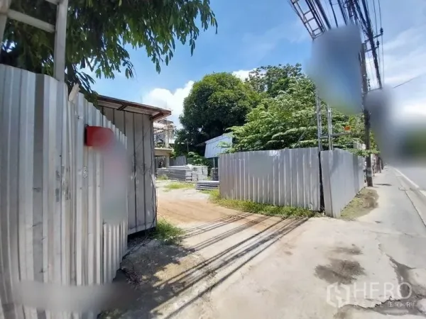 Land for rent in Khlong Toei, Bangkok - Gated land plot with corrugated metal fence, dirt driveway, and roadside access in Khlong Toei under a blue sky.