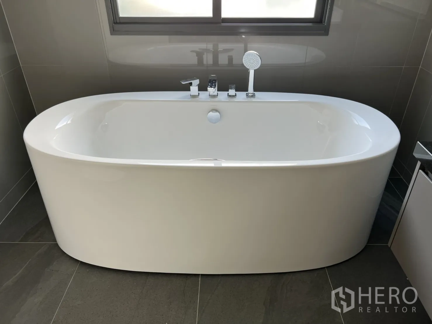 Detached House for rent in Bang Kapi, Bangkok - Bathroom featuring a freestanding oval bathtub beneath a window and gray tiles.