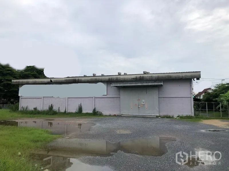 Warehouse for rent in Bang Kapi, Bangkok - Single-storey warehouse exterior with large steel doors, gravel driveway and open yard.