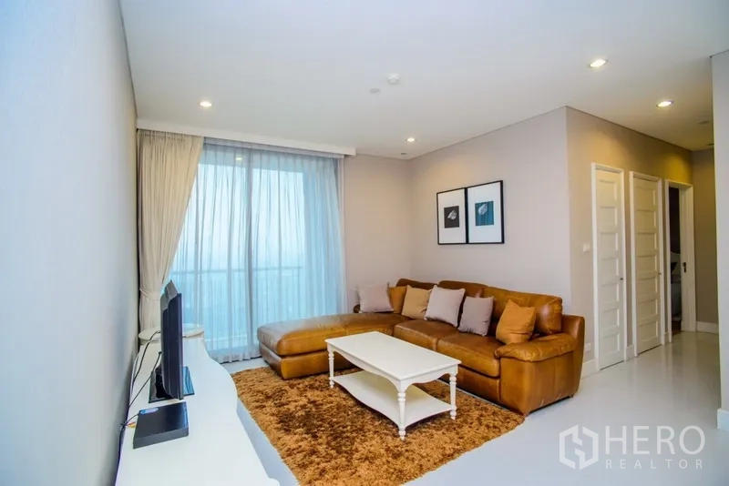 Condominium for rent in Khlong Toei, Bangkok - Bright living room with a leather sectional sofa, white coffee table, TV console and floor-to-ceiling window to a balcony view.